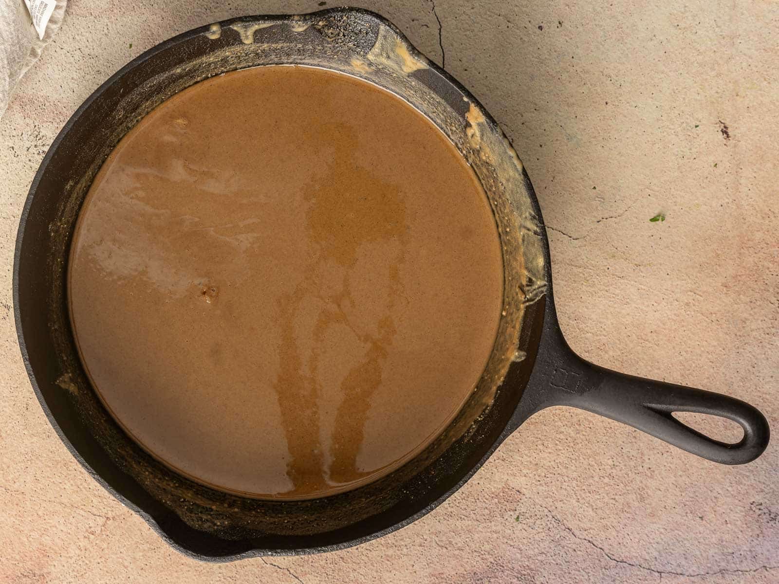 A cast iron skillet brims with a rich, gluten-free roux, its brown liquid batter resting elegantly on a textured surface.