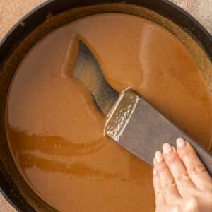 Hand stirring a rich, gluten-free roux in a black skillet with a flat wooden spatula, perfect for adding depth to your gumbo.
