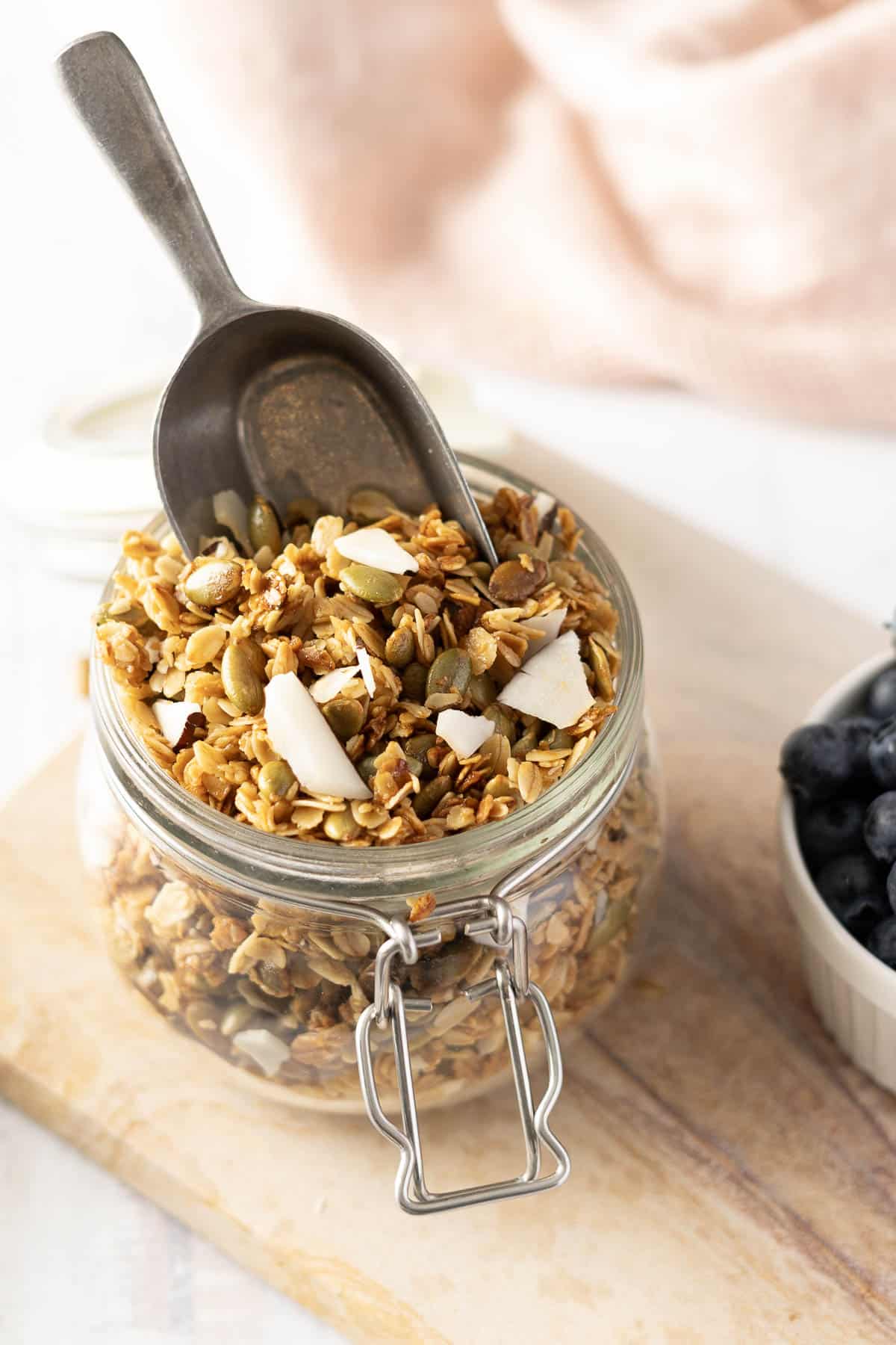 A glass jar filled with honey granola and a metal scoop on a wooden board, next to a bowl of blueberries.