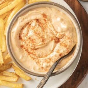 A bowl of creamy aioli sprinkled with paprika sits next to a side of fries, offering a hint of spicy indulgence. A spoon rests invitingly in the bowl.