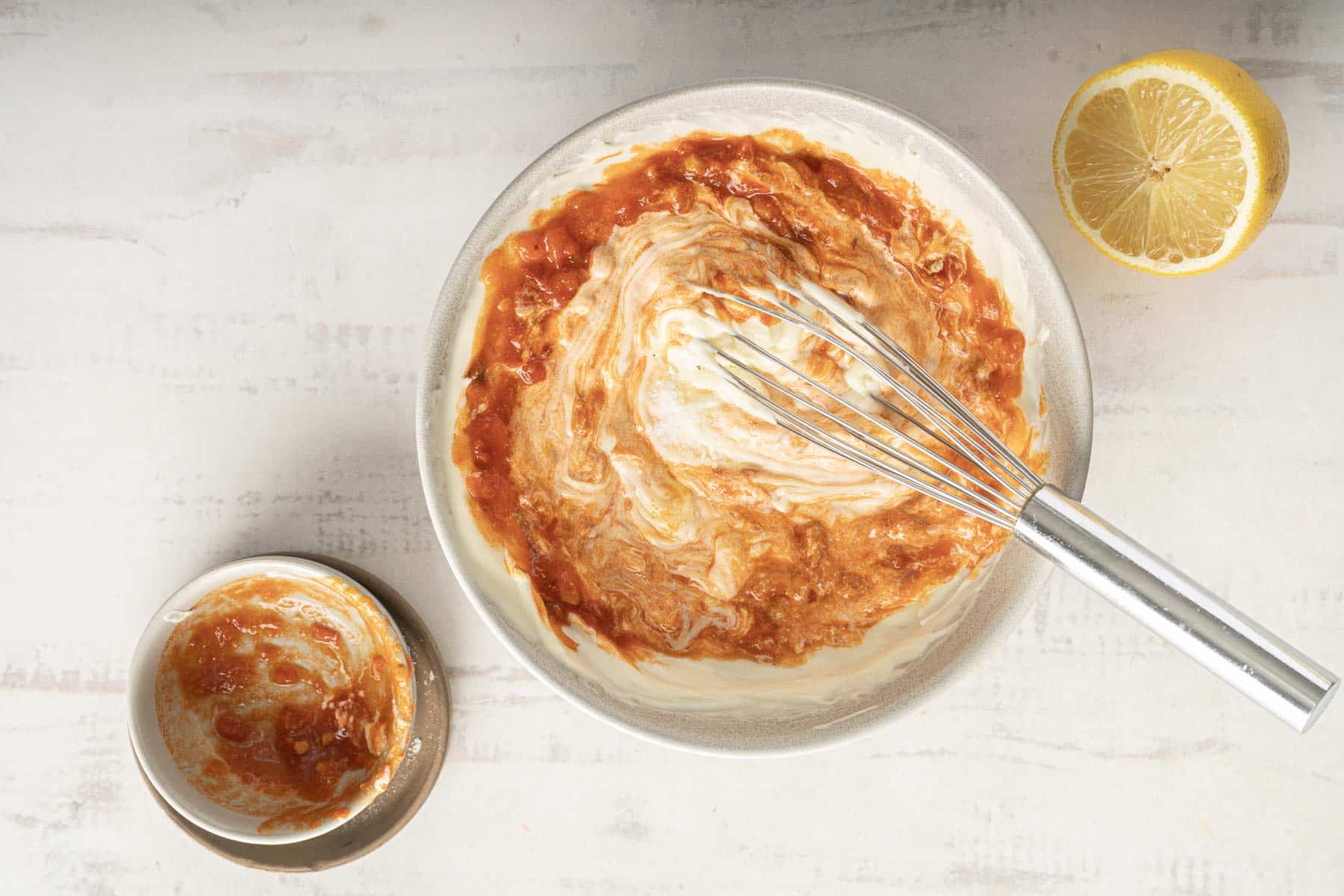 A whisk blends yogurt with spicy harissa aioli in a bowl, while a small dish of the vibrant harissa and half a lemon rest nearby.