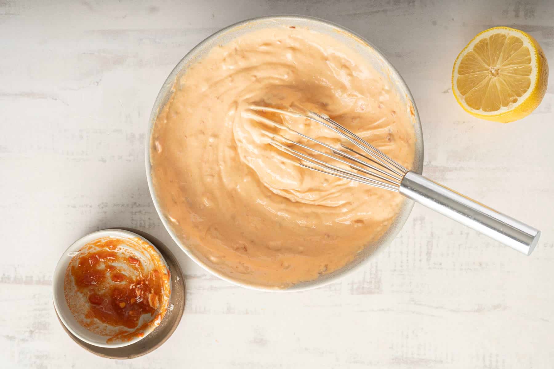 A bowl of creamy aioli with a whisk sits next to a halved lemon and a small dish filled with thick, spicy harissa, all on a pristine white surface.
