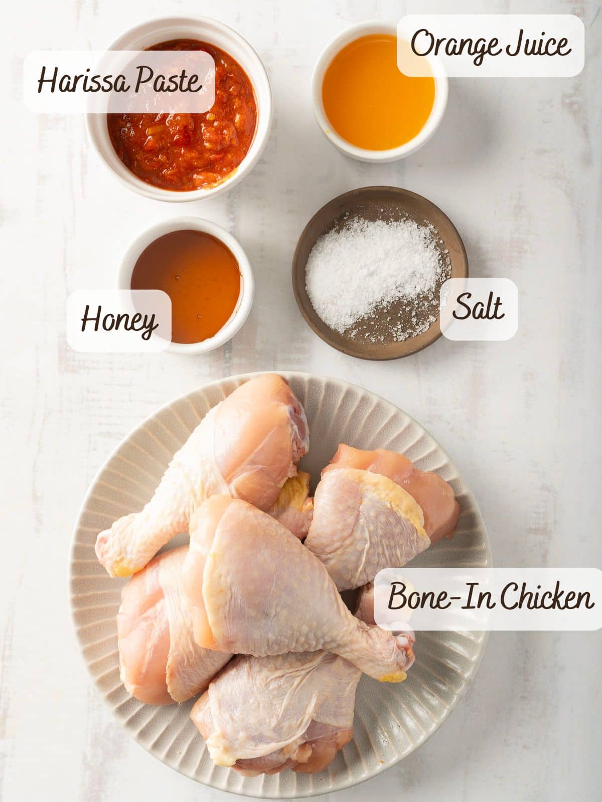 A plate with bone-in chicken legs and bowls containing harissa paste, orange juice, honey, and salt are arranged on a white surface, ready to create a delightful Honey Harissa Chicken.