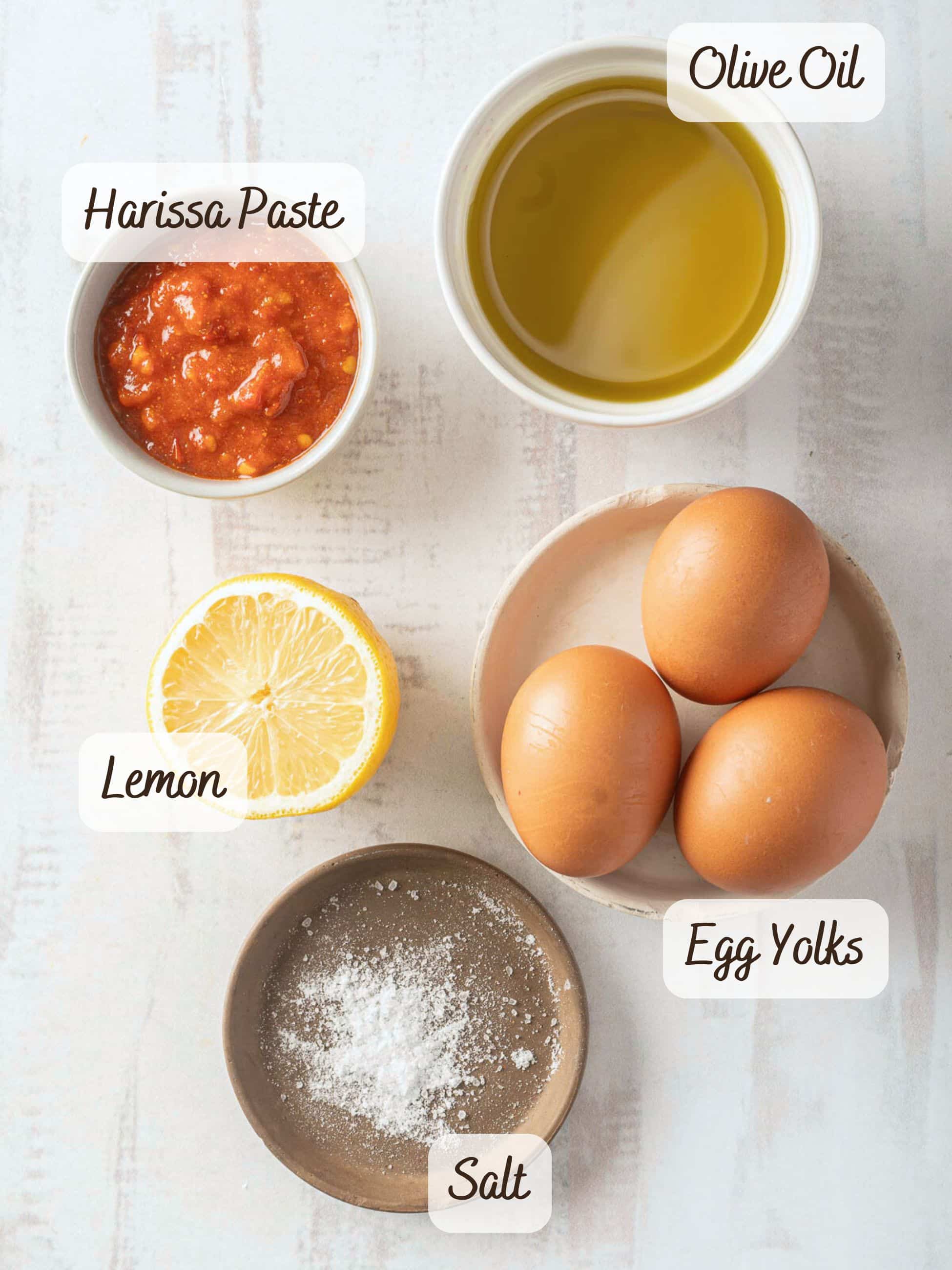 Ingredients displayed: olive oil in a bowl, three egg yolks, half a lemon, harissa paste in a bowl for a spicy kick, and a dish of salt on a light surface. Perfect for crafting a zesty aioli with flair.