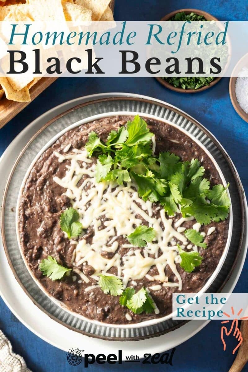 A bowl of homemade refried black beans topped with melted cheese and cilantro on a plate, with tortilla chips on the side.