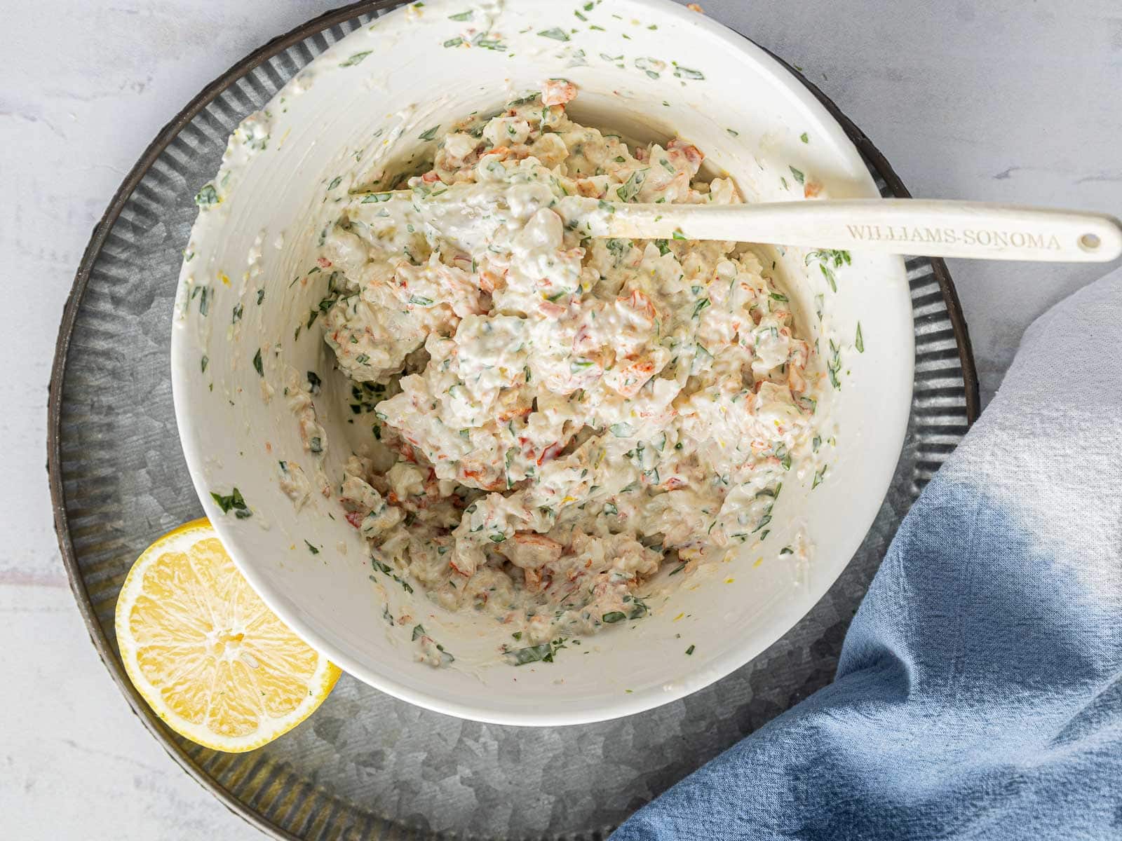 A bowl of crab and lobster salad with a wooden spoon, garnished with herbs, next to a half lemon and a blue napkin on a textured surface.