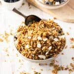 A bowl of honey granola with coconut flakes and seeds, placed on a white surface with a spoon inside.