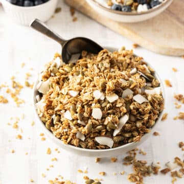 A bowl of honey granola with coconut flakes and seeds, placed on a white surface with a spoon inside.