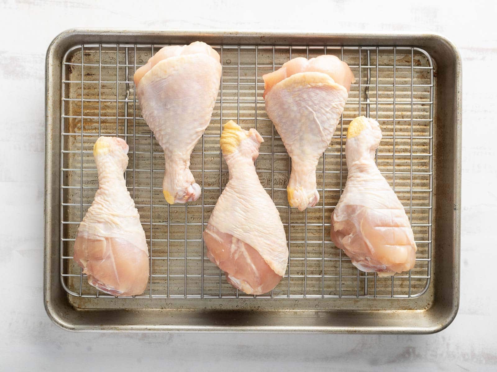 Five raw chicken drumsticks, glazed with a hint of harissa and honey, are artfully arranged on a wire rack placed over a baking sheet.