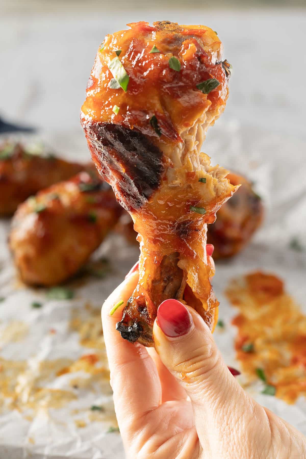 A hand holding a grilled chicken drumstick coated in a sticky honey sauce, with more succulent chicken legs glistening on foil in the background.
