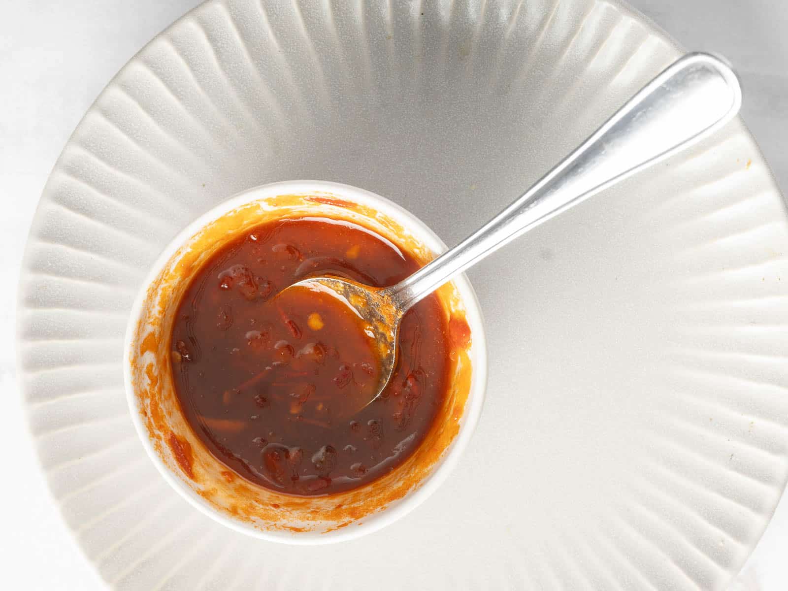 A small white bowl filled with red sauce containing visible chunks and a hint of harissa, placed on a textured plate. A metal spoon is resting inside the bowl.