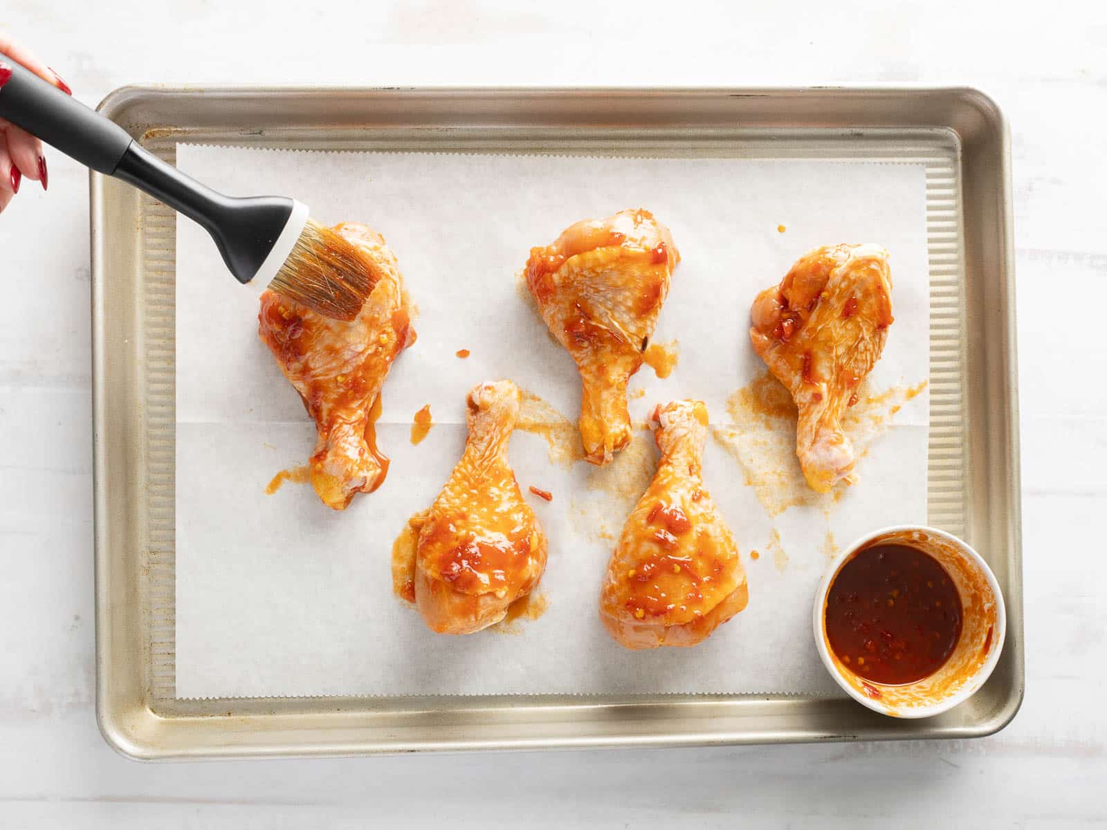 Brushing a zesty honey harissa marinade on five chicken drumsticks laid out on a parchment-lined baking sheet.