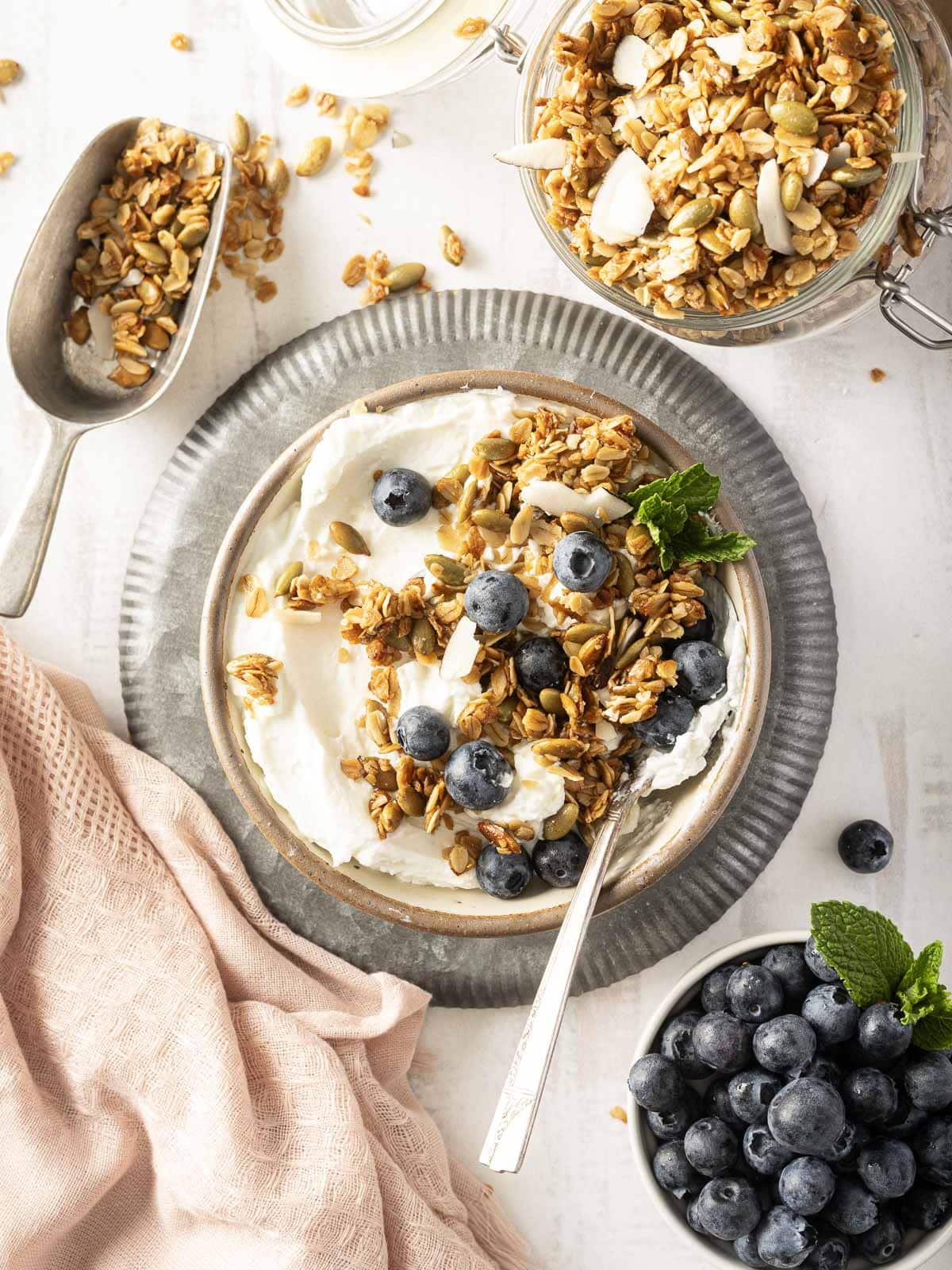 A bowl of yogurt topped with honey granola and blueberries sits ready with a spoon. Nearby, a jar and a scoop of granola, alongside a bowl of blueberries, rest on a pink cloth.