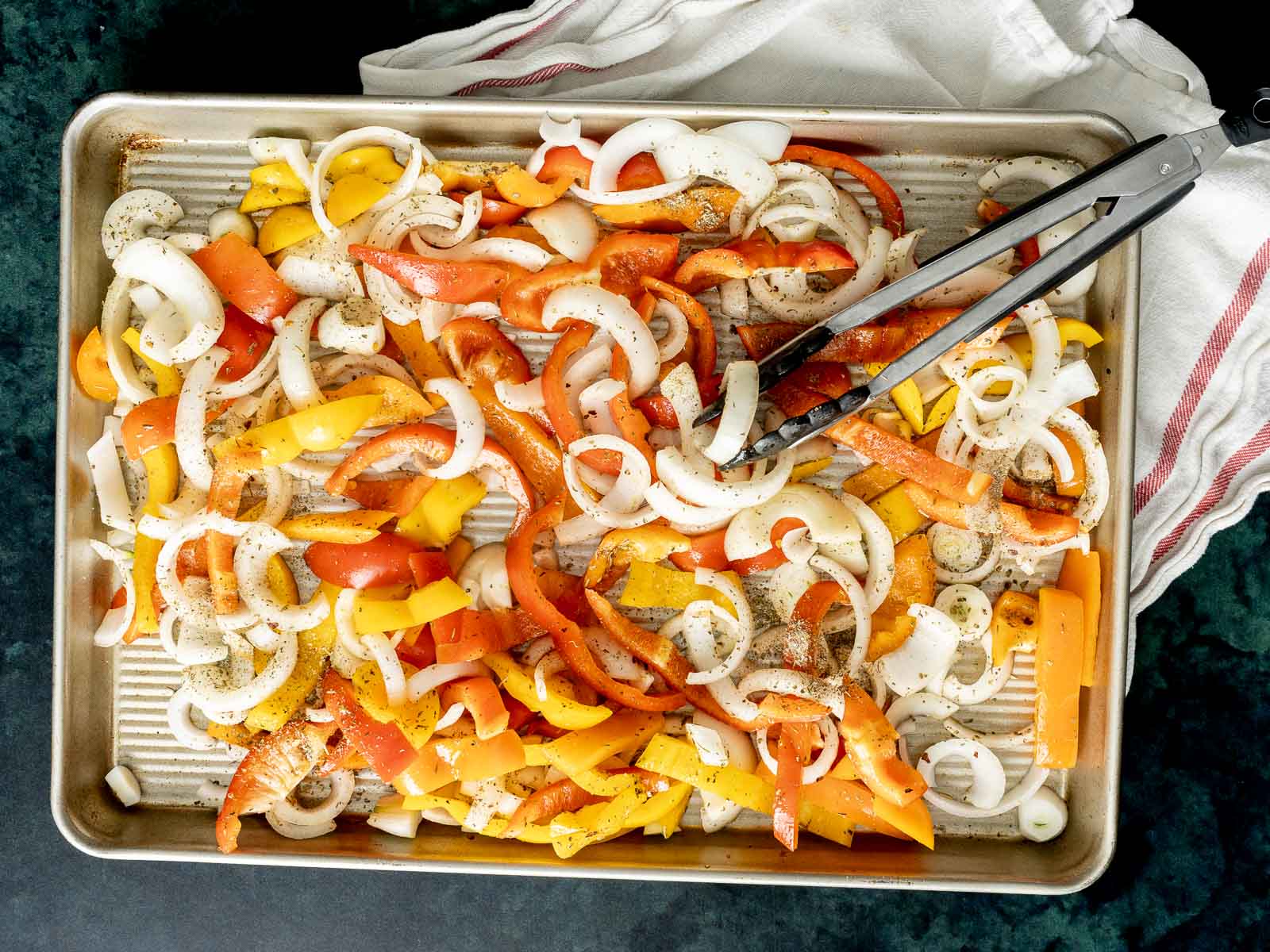 A baking sheet filled with sliced yellow and red bell peppers, onions, savory baked Italian sausage, and seasoning, with metal tongs and a cloth in the corner.