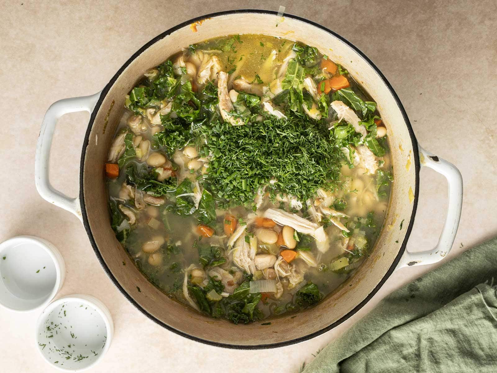 A pot of hearty chicken and white bean soup with kale, carrots, and herbs sits on a light-colored surface. Nearby, two small bowls and a green cloth complete the cozy scene.