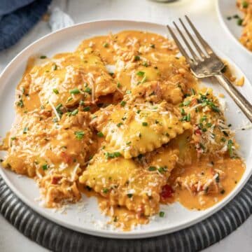 A plate of lobster ravioli with creamy tomato sauce, topped with grated cheese and chopped chives, sits invitingly next to a fork on a white plate.