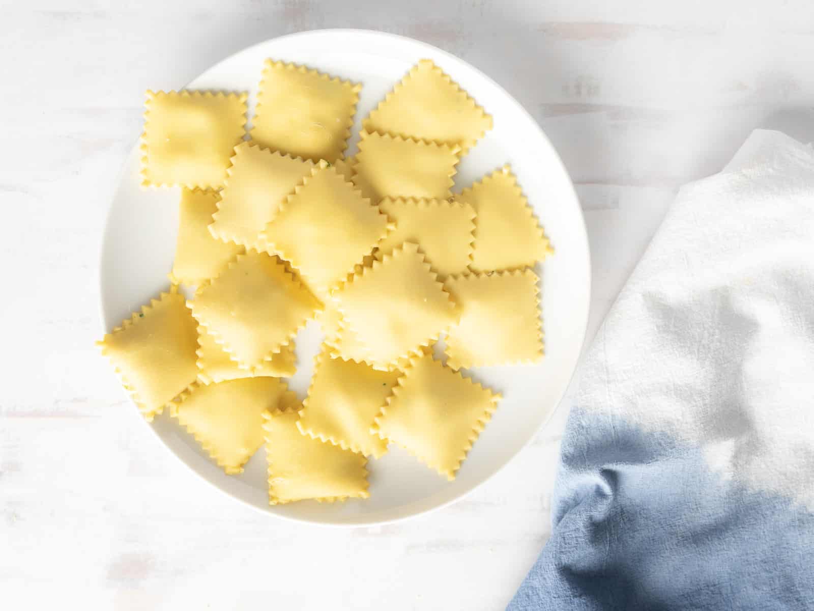 A white plate with uncooked ravioli squares, potentially filled with delectable lobster, rests next to a white and blue cloth on a light surface.