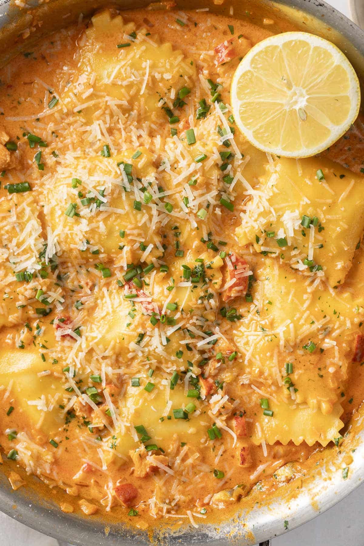 A skillet of lobster ravioli in a creamy tomato sauce topped with grated cheese and chopped herbs, garnished with a lemon slice.