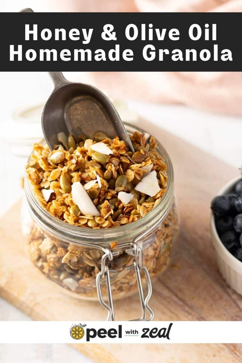 A jar of homemade granola with a metal scoop on a wooden surface, next to a bowl of blueberries.