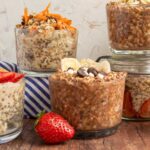 A variety of glass jars filled with overnight oats, topped with fruit, chocolate chips, and nuts. A fresh strawberry is placed in the foreground.