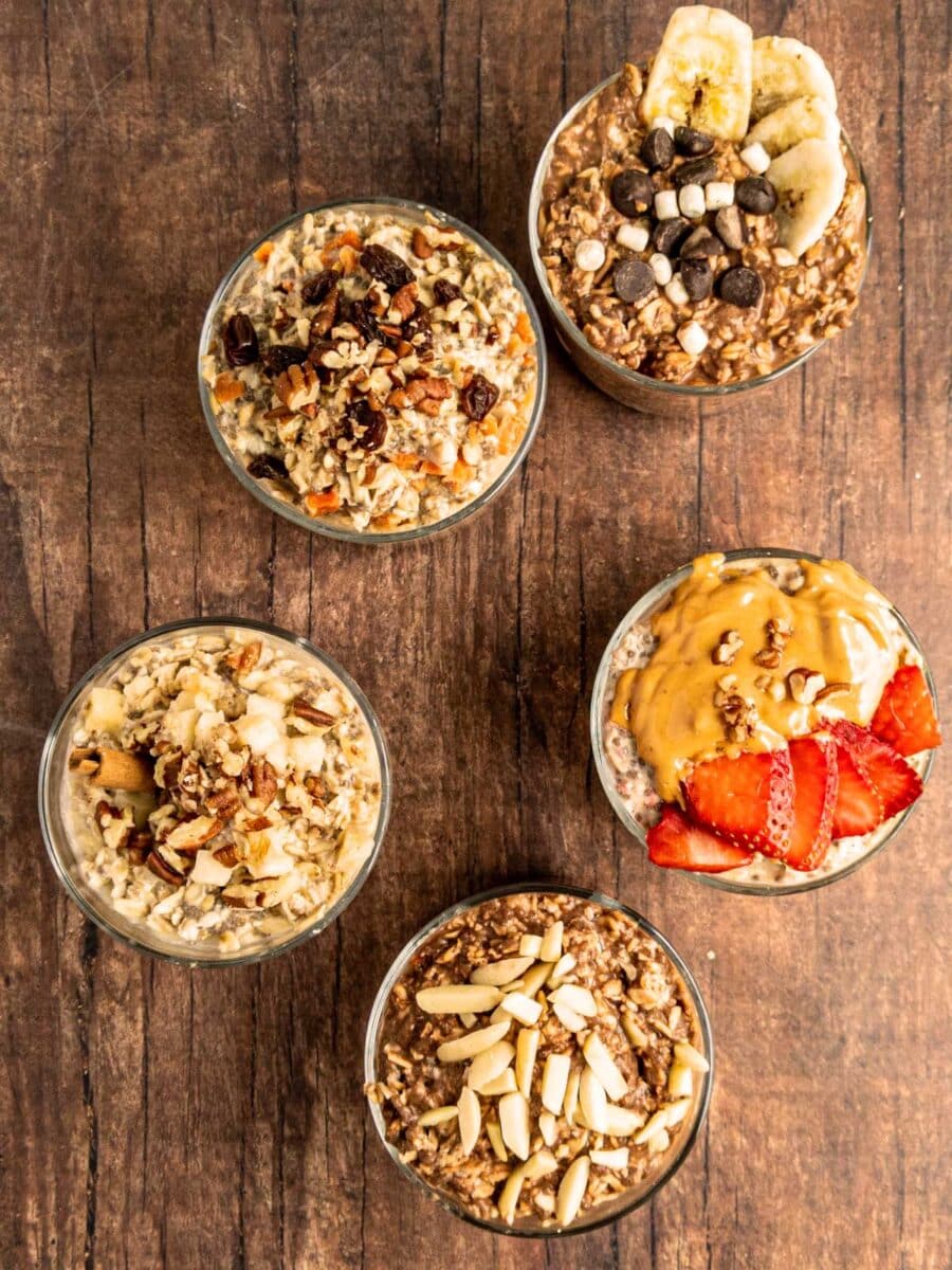 Five glass cups with various oatmeal toppings including nuts, chocolate chips, strawberries, bananas, and dried fruits arranged on a wooden surface.