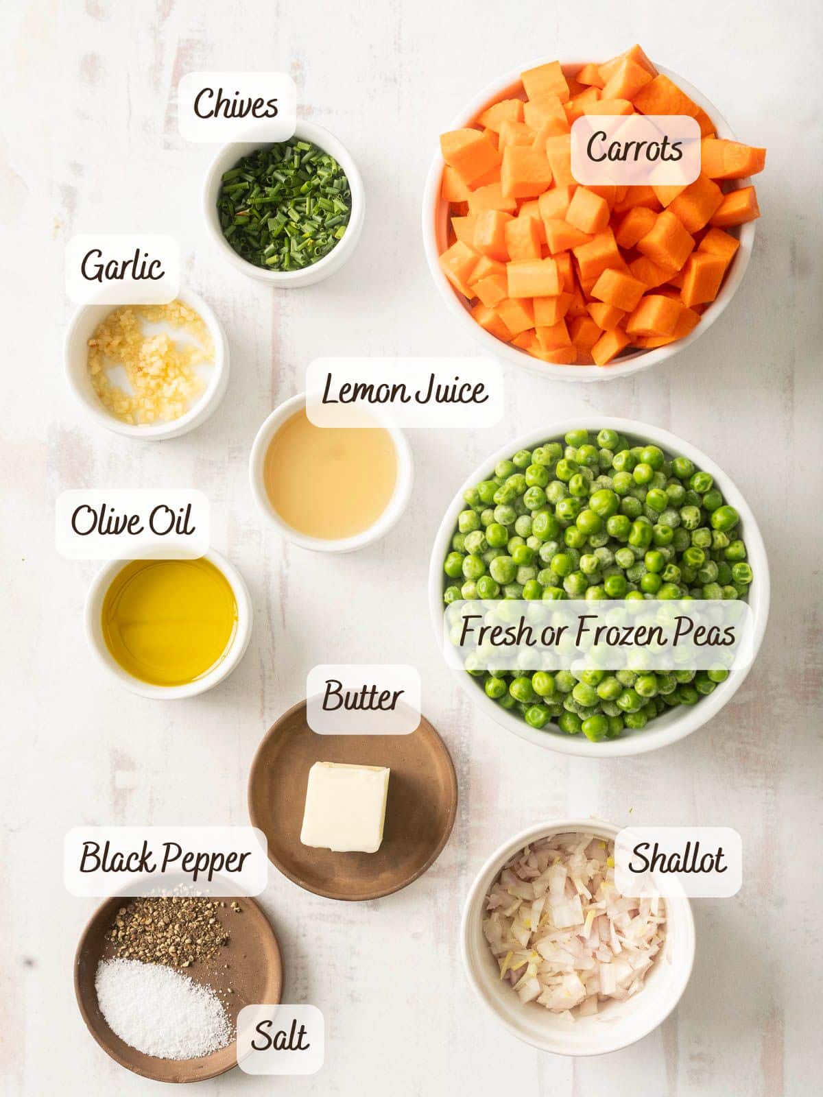 Bowls of ingredients are arranged on a white surface: chopped carrots and peas, chives, minced garlic, lemon juice, olive oil, butter, chopped shallot, black pepper, and salt.