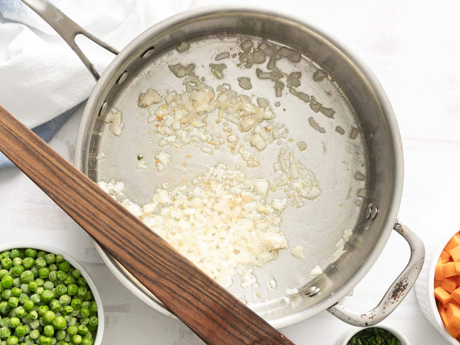 Chopped onions sautéing in a stainless steel pan, alongside bowls brimming with fresh peas and carrots, with a wooden spatula resting on the pan's edge.