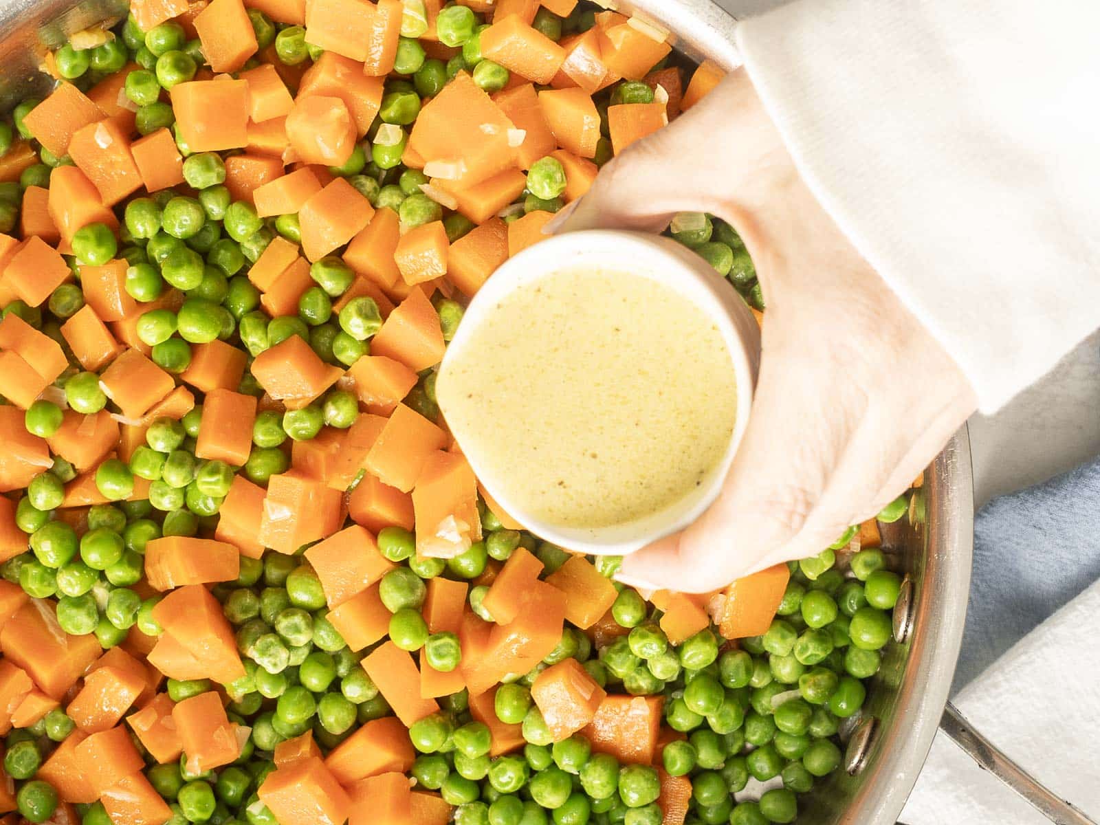 A hand hovers above a pan filled with vibrant peas and carrots, gently sprinkling a bowl of seasoning to enhance the flavors.