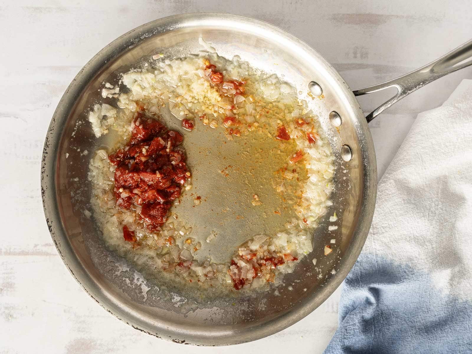 A metal frying pan sizzles with chopped onions, minced garlic, and sun-dried tomatoes cooking in oil, setting the stage for a delightful tomato cream sauce. A white and blue cloth is partially visible on the side, waiting to serve up some succulent lobster ravioli.