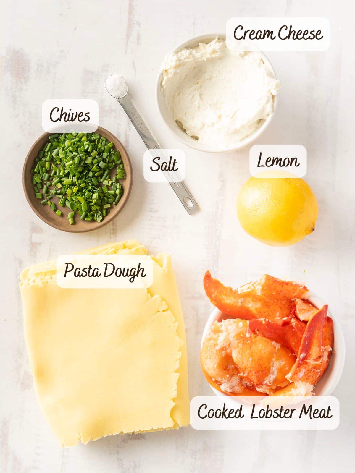 Ingredients laid out on a table: cream cheese in a bowl, chopped chives on a plate, a measuring spoon of salt, a lemon, pasta dough sheet, and a bowl of cooked lobster meat ready to be wrapped into delicate seafood ravioli.