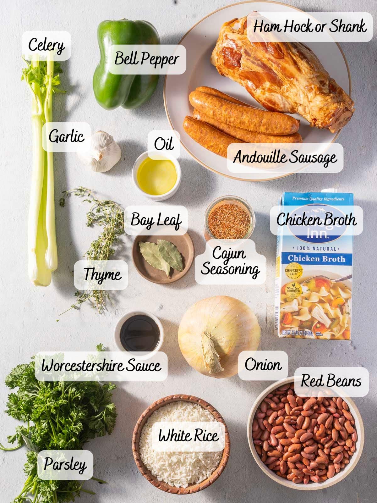 Ingredients for a Cajun dish are arranged: celery, bell pepper, ham hock, andouille sausage, garlic, oil, bay leaf, thyme, chicken broth, onion, red beans, Worcestershire sauce, rice, and parsley.