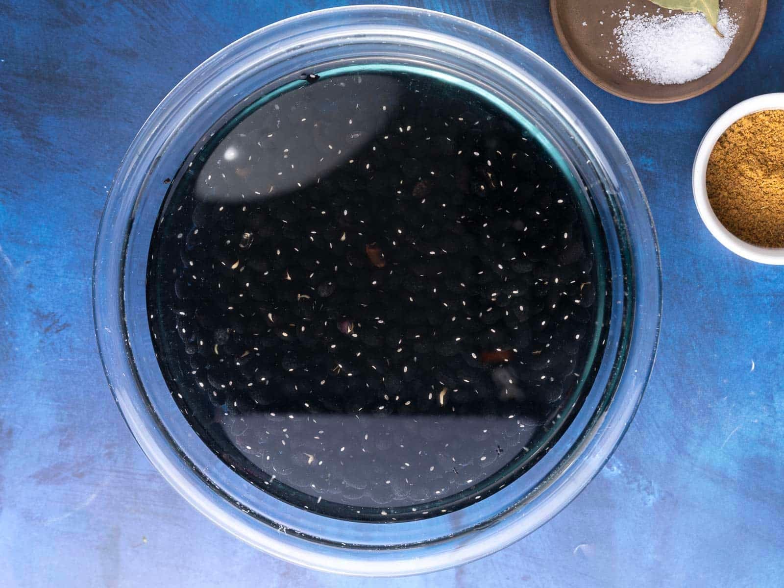 A bowl of black beans soaking in water on a blue surface, with small bowls close by.
