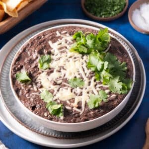 A bowl of refried black beans is topped with melted cheese and cilantro, placed on a stack of plates.