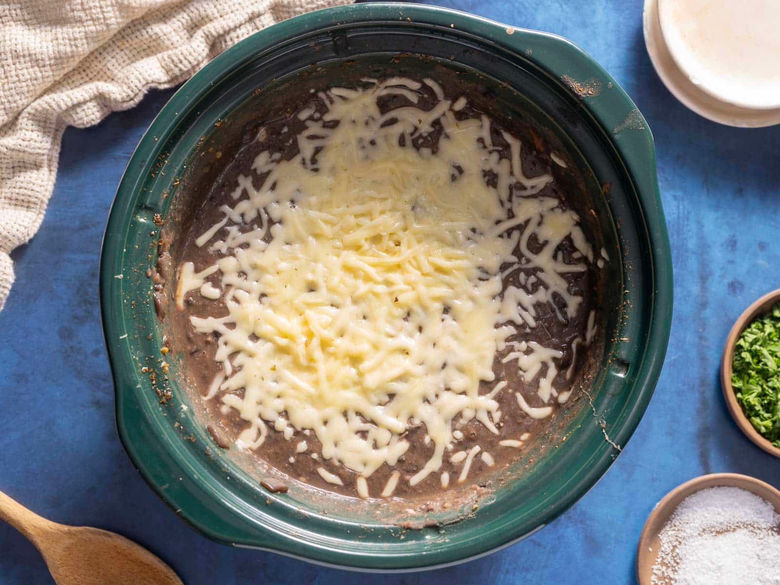A crockpot filled with  refried black beans, topped with melted cheese, surrounded by a cloth, wooden spoon, and small bowls of chopped herbs and salt on a blue surface.