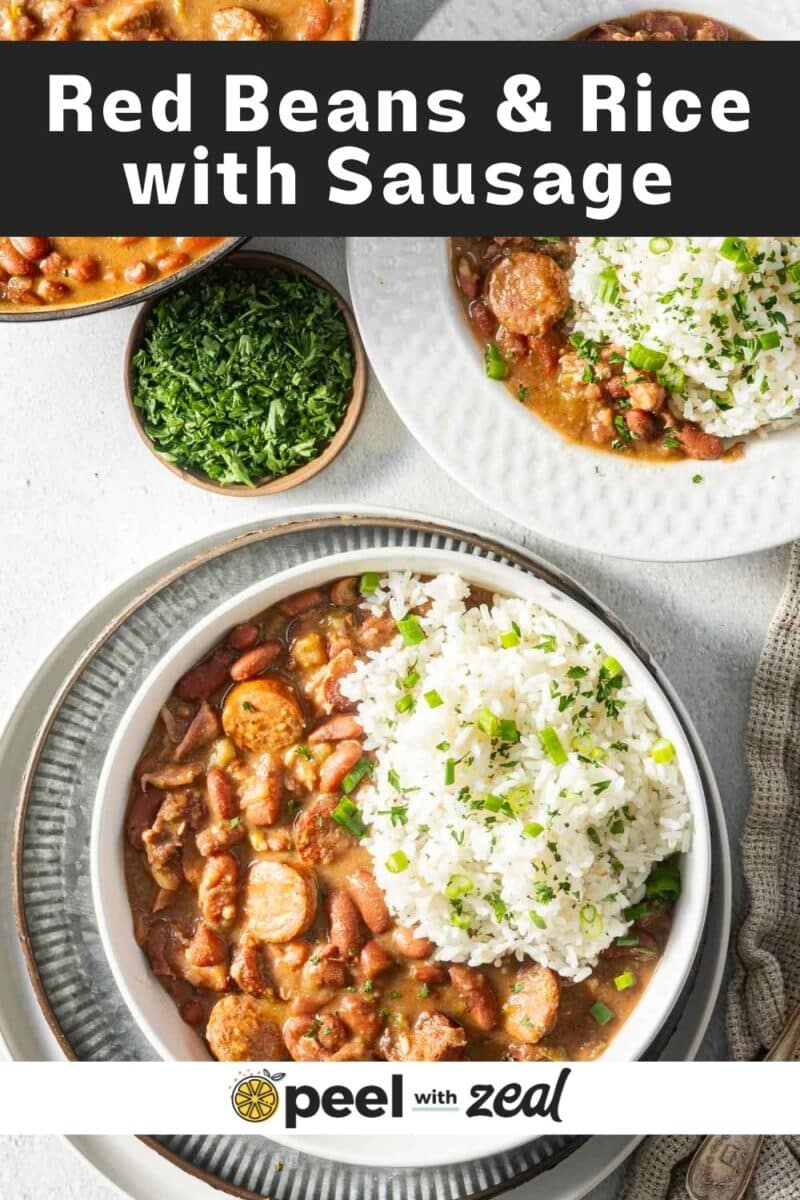 A bowl of red beans, sausage, and white rice garnished with chopped herbs, with a side bowl of herbs, on a textured surface.