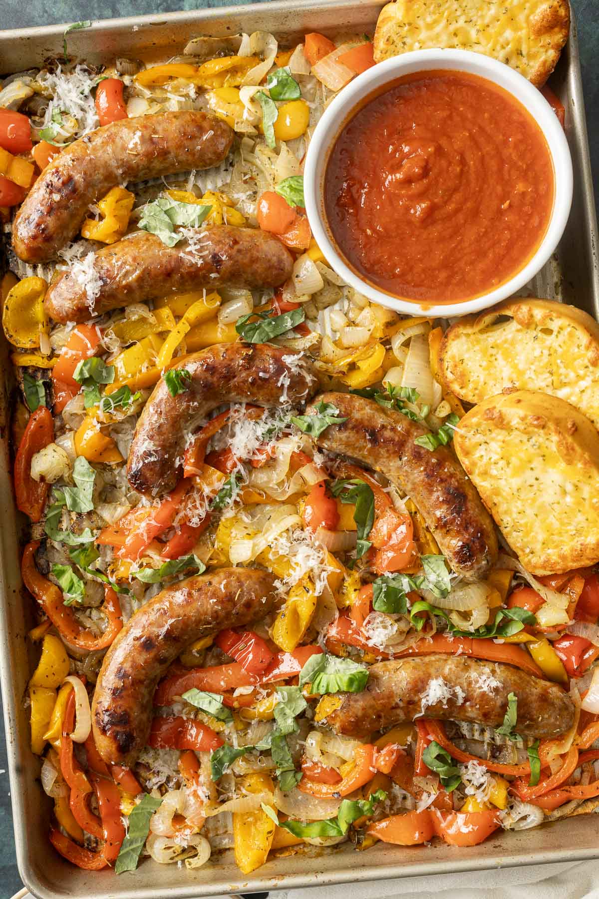 Baked Italian sausage nestled on a sheet pan with roasted bell peppers, onions, and fragrant herbs, served alongside a bowl of red sauce and slices of toasted bread.