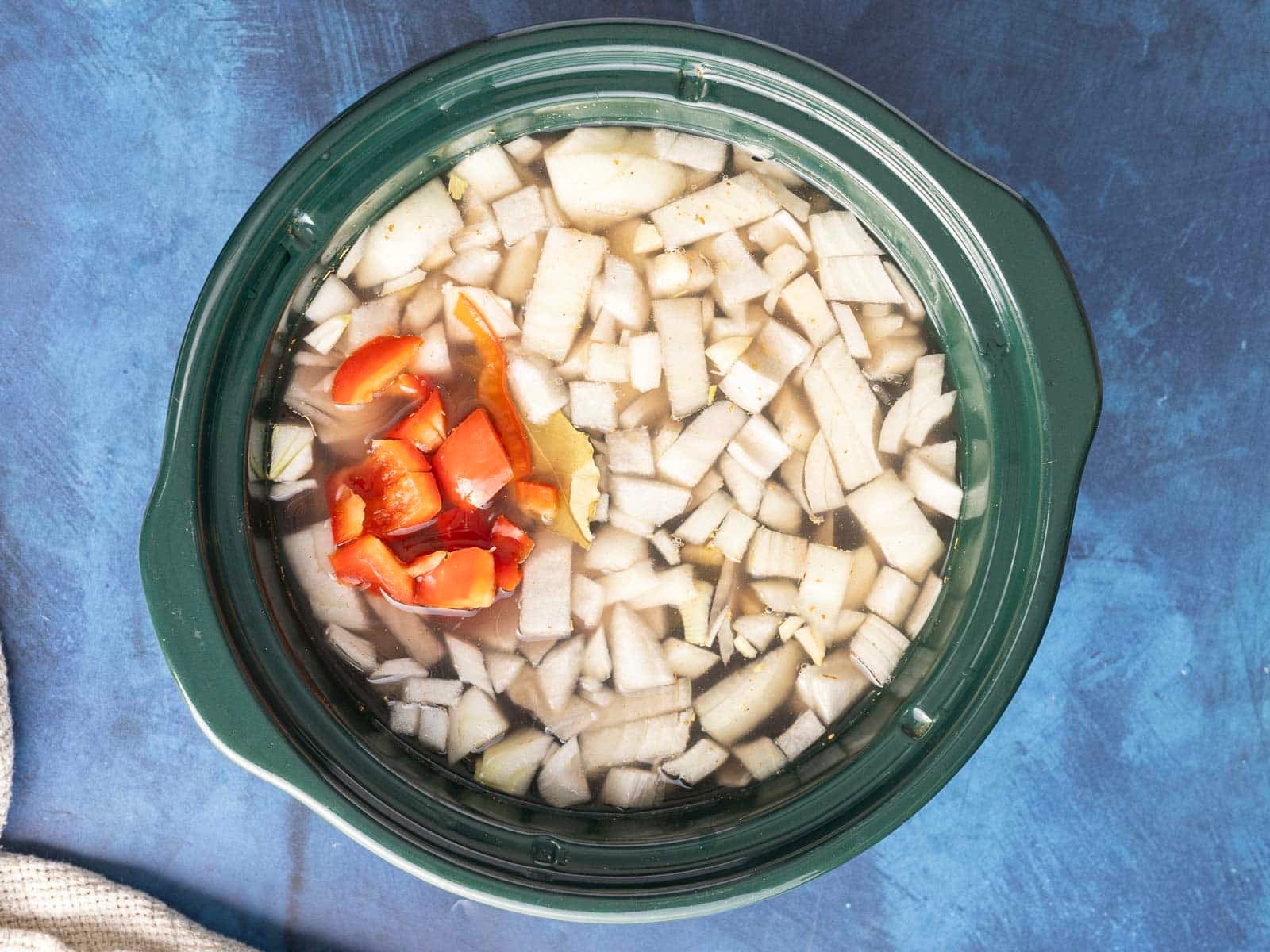 A slow cooker brimming with diced onions, red peppers, black beans, and a bay leaf in liquid rests on a blue surface.