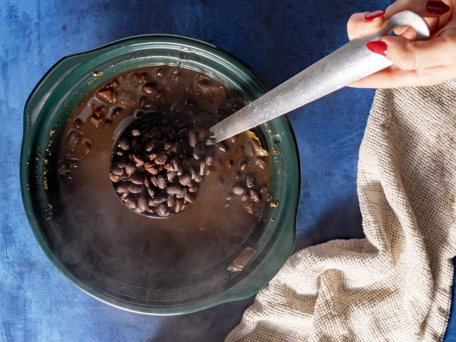 A person ladles black beans from a slow cooker on a blue surface. Steam rises from the pot, and a beige cloth is nearby.