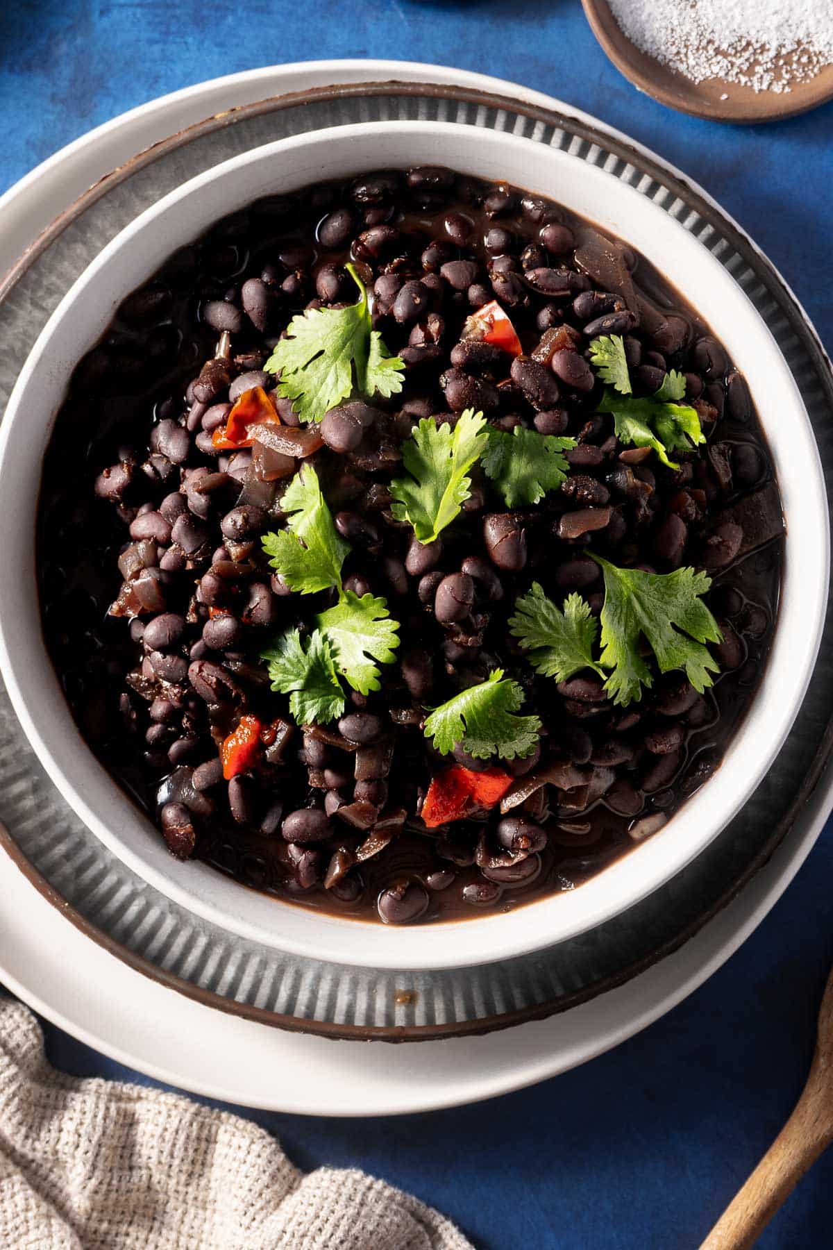 A bowl of slow-cooked black beans, garnished with cilantro and small red pepper pieces, sits invitingly on a textured blue tabletop, perfect for trying out a new recipe.