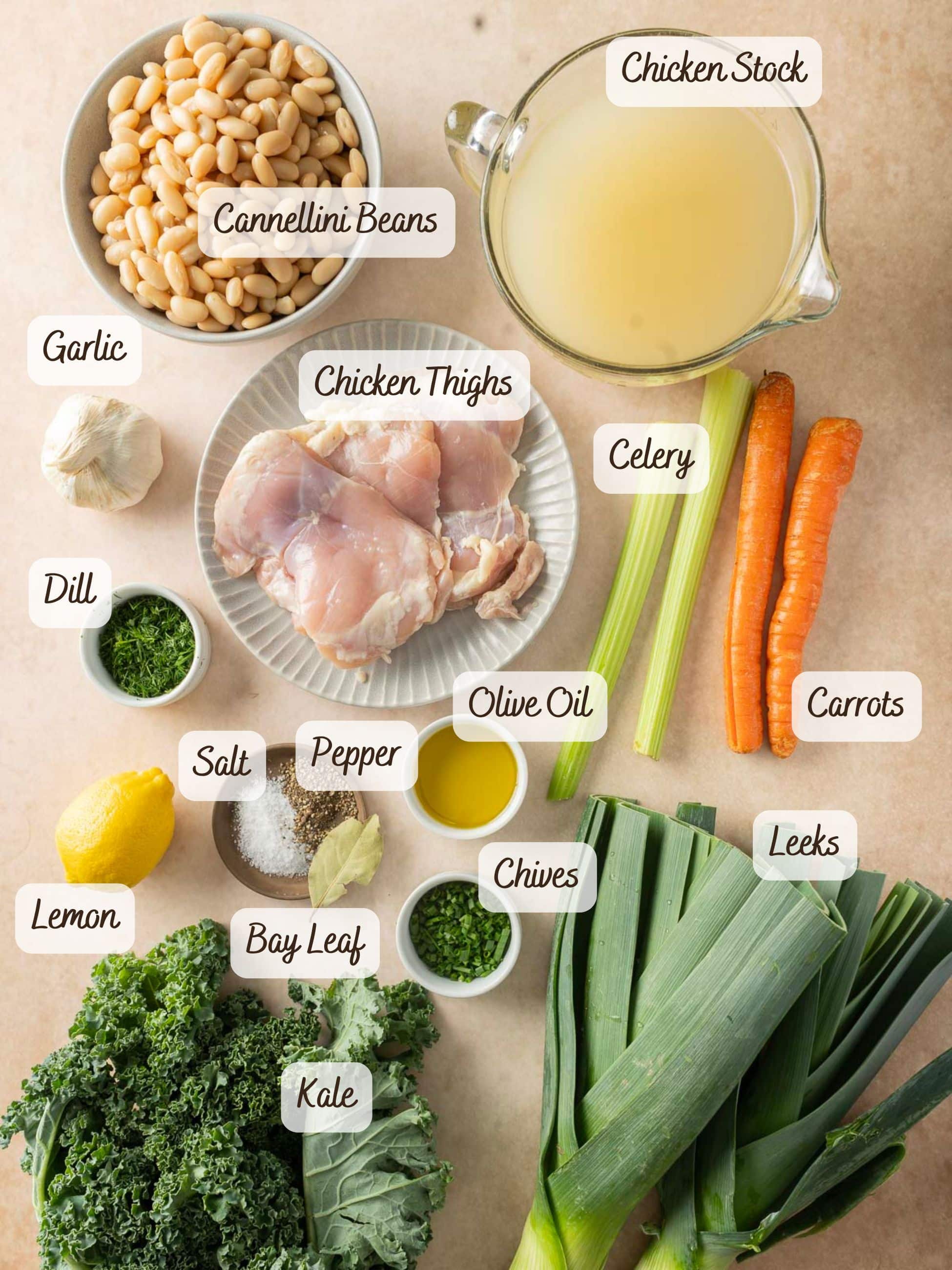 Ingredients neatly arranged for a hearty white bean soup: cannellini beans, chicken stock, chicken thighs, celery, carrots, garlic, dill, olive oil, chives, kale, leeks, lemon, salt, pepper, and a bay leaf. Perfectly crafted for a comforting meal.