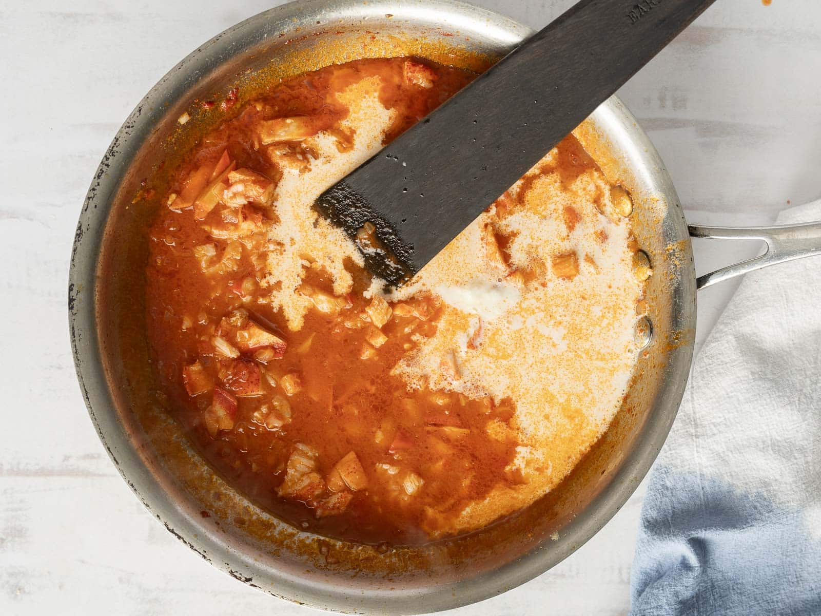 Simmering stew in a stainless steel pan with a spatula resting on the edge; chunks of meat and a creamy tomato cream sauce visible.