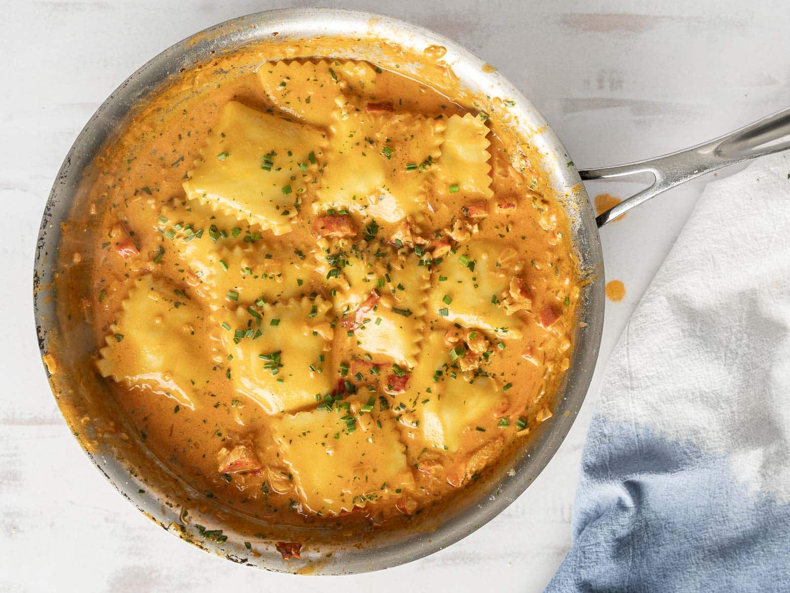 A pan filled with lobster ravioli in a rich tomato cream sauce, garnished with herbs. A cloth is partially visible on the side.