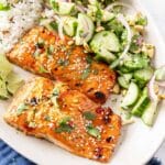 Plate of sweet chili salmon garnished with sesame seeds and cilantro, served alongside rice and a refreshing cucumber salad.