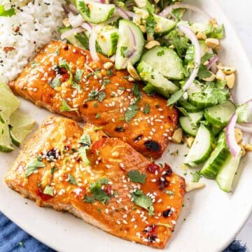Plate of sweet chili salmon garnished with sesame seeds and cilantro, served alongside rice and a refreshing cucumber salad.