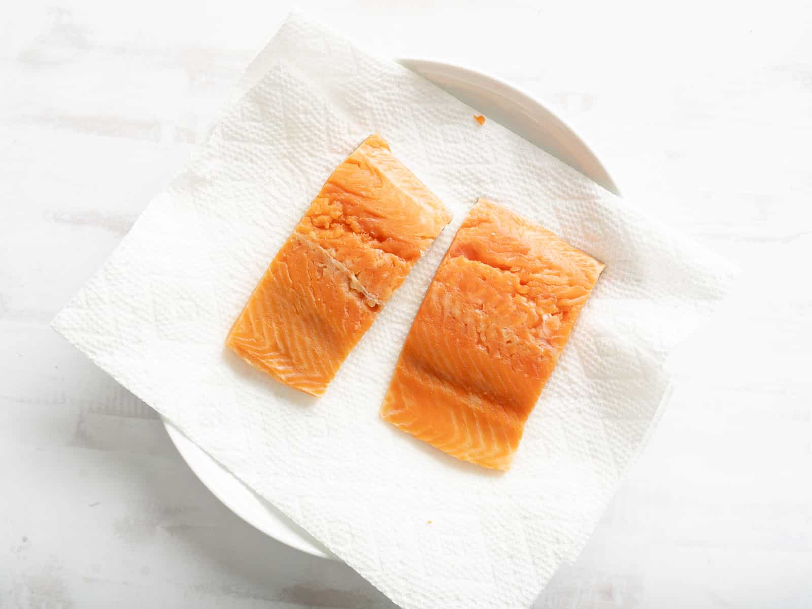 Two raw salmon fillets rest on a white paper towel covering a plate.