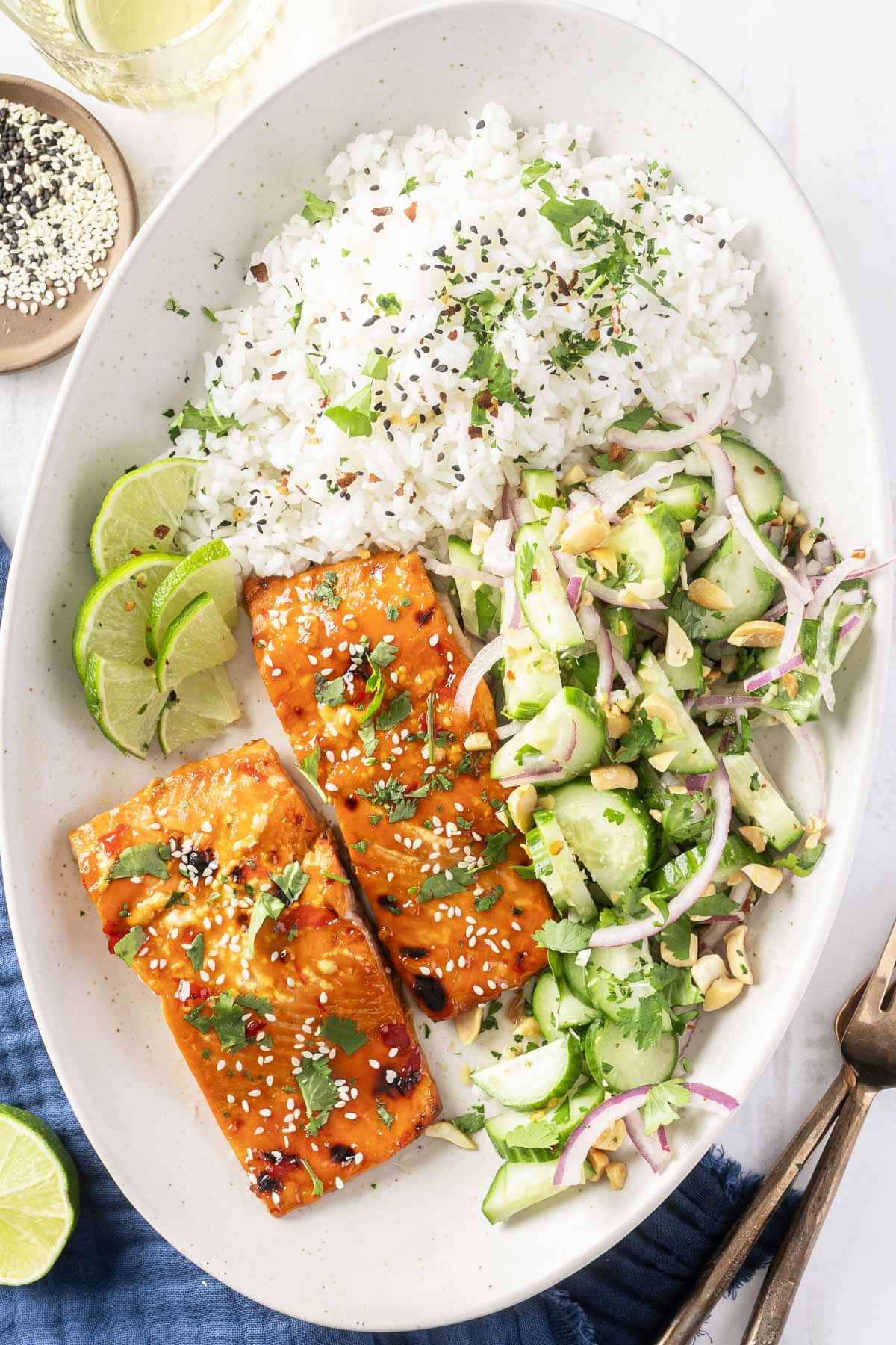 A plate showcasing a sweet chili salmon recipe, featuring sesame-glazed salmon paired with white rice garnished with herbs, a cucumber-avocado salad, and lime wedges.