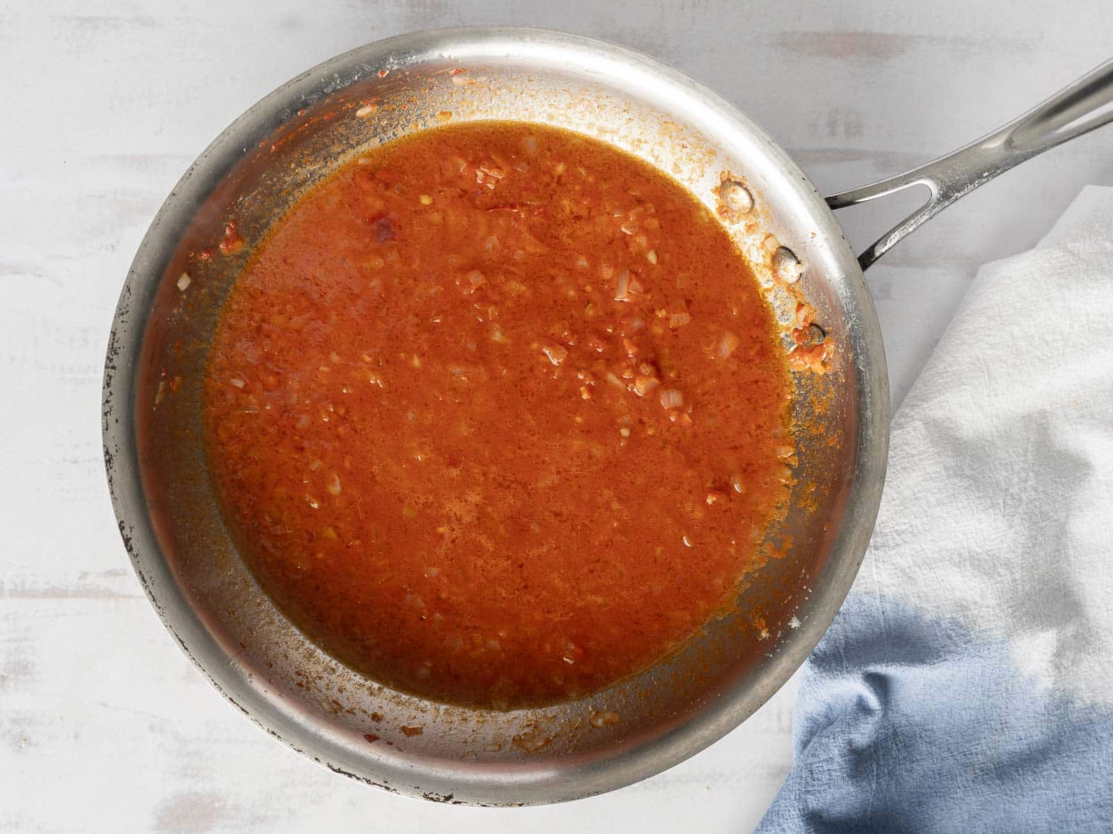 A stainless steel pan with a reddish-brown Lobster Ravioli sauce containing small onion pieces. A white and blue cloth is beside the pan on a light-colored surface, hinting at the delicious meal soon to be enjoyed.