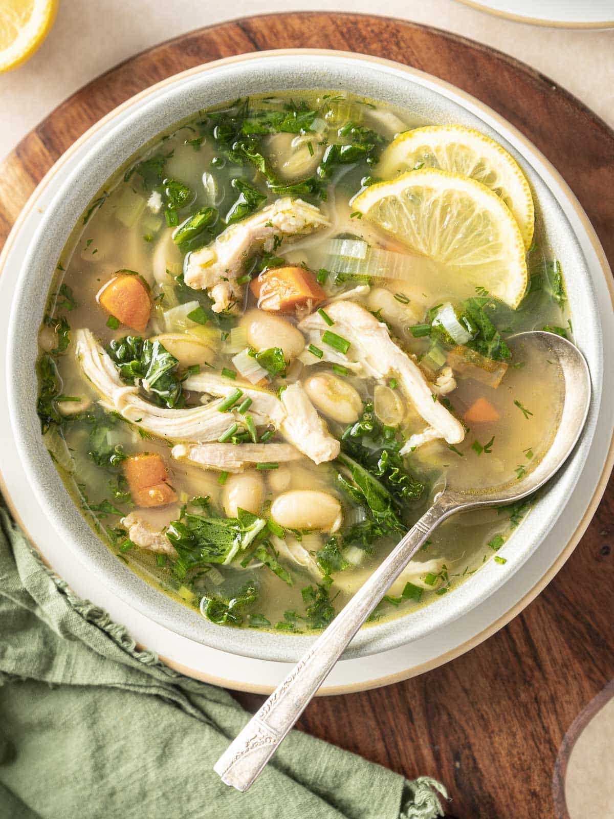 A hearty bowl of chicken soup with white beans, kale, carrot chunks, and lemon slices is served with a spoon on a rustic wooden board.