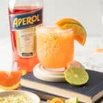 A glass of Aperol margarita with lime and orange slices, next to an Aperol bottle, rests elegantly on a stack of books atop a marble surface, evoking the charm of a classic Margarita in its vibrant presentation.