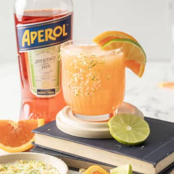 A glass of Aperol margarita with lime and orange slices, next to an Aperol bottle, rests elegantly on a stack of books atop a marble surface, evoking the charm of a classic Margarita in its vibrant presentation.